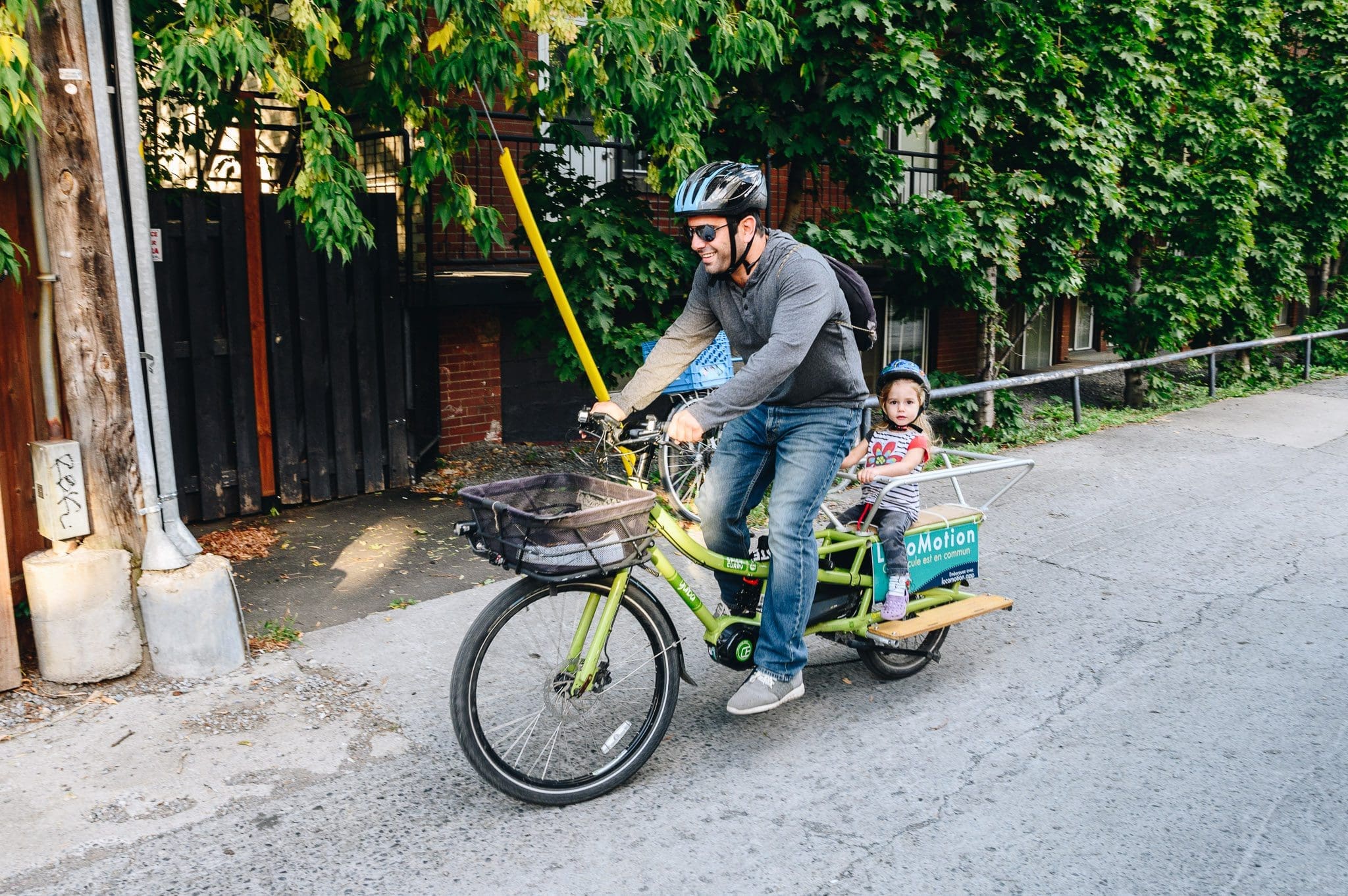 Un homme portant un casque pédale sur un vélo cargo vert dans la rue, son jeune enfant assis derrière lui, tandis que des bâtiments couverts de lierre défilent. Le vélo est équipé d'un panier arrière très pratique. Audrey McMahon a capturé cette charmante scène urbaine.