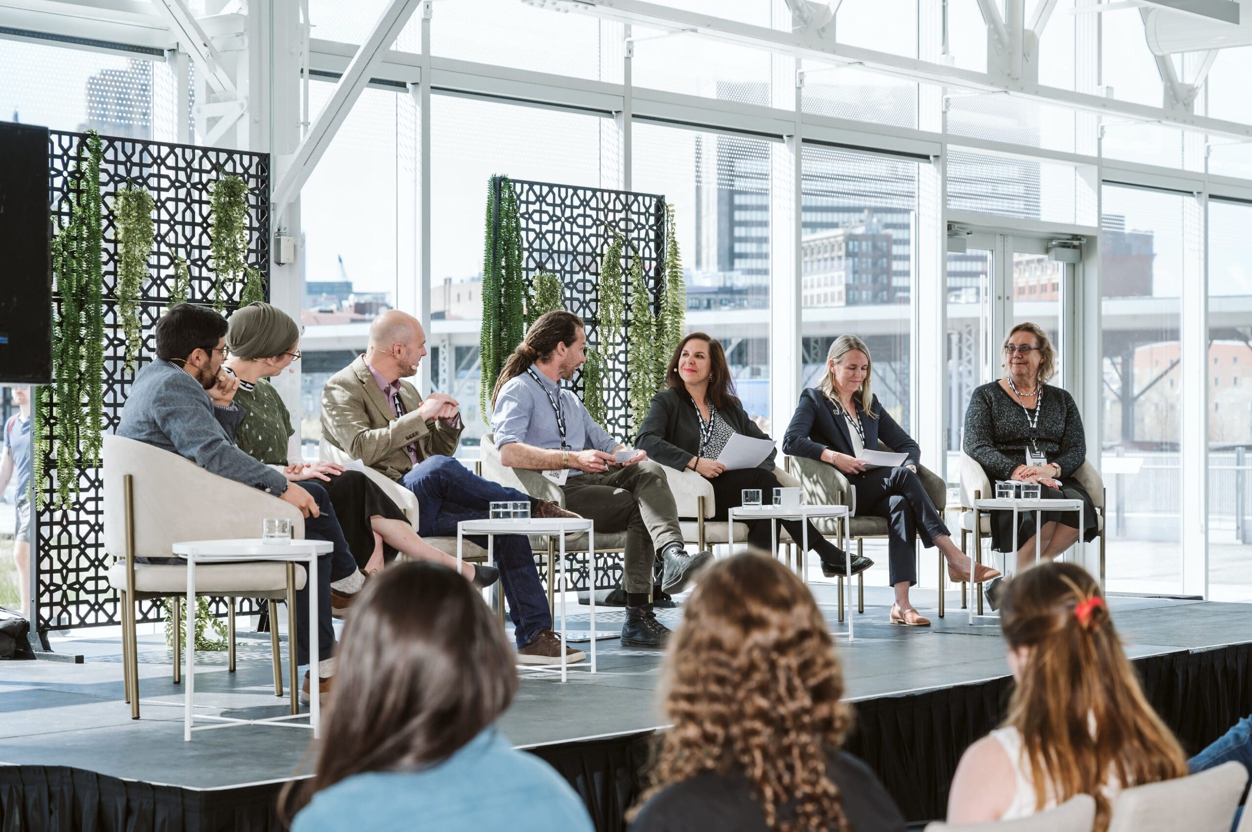 Un groupe de six personnes, hommes et femmes, est assis sur une scène du Sommet Climat 2024, avec des planchettes et des microphones, et participe à une discussion. Deux membres du public sont visibles au premier plan. Le lieu est moderne, avec de grandes fenêtres montrant un paysage urbain.