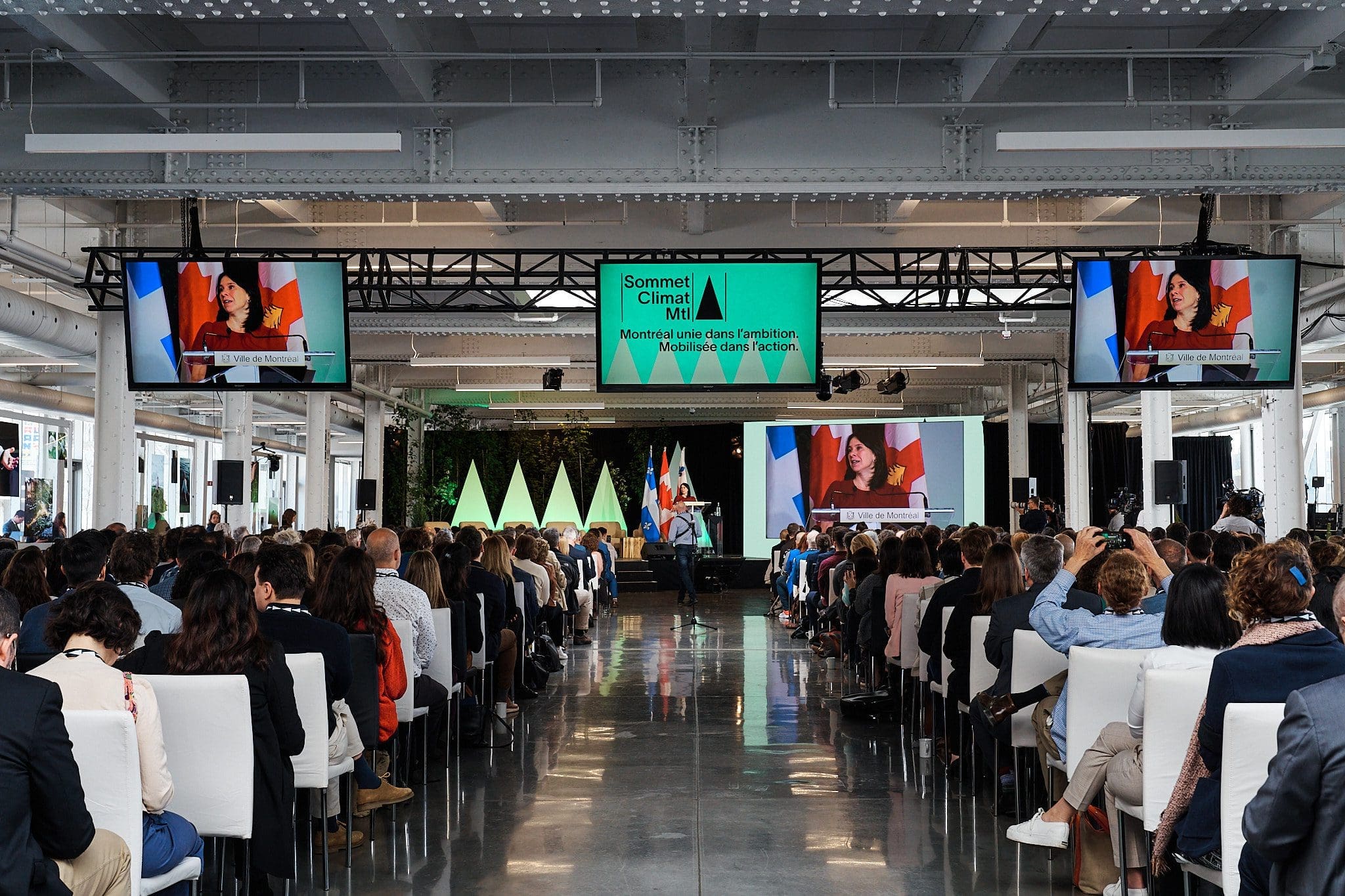 Un large public est assis en rangs face à une scène lors d'une conférence du Sommet Climat 2023. Sur la scène se trouvent un orateur sur un podium, plusieurs écrans et des bannières. La salle est bien éclairée par de grandes fenêtres et plusieurs écrans montrent l'orateur.