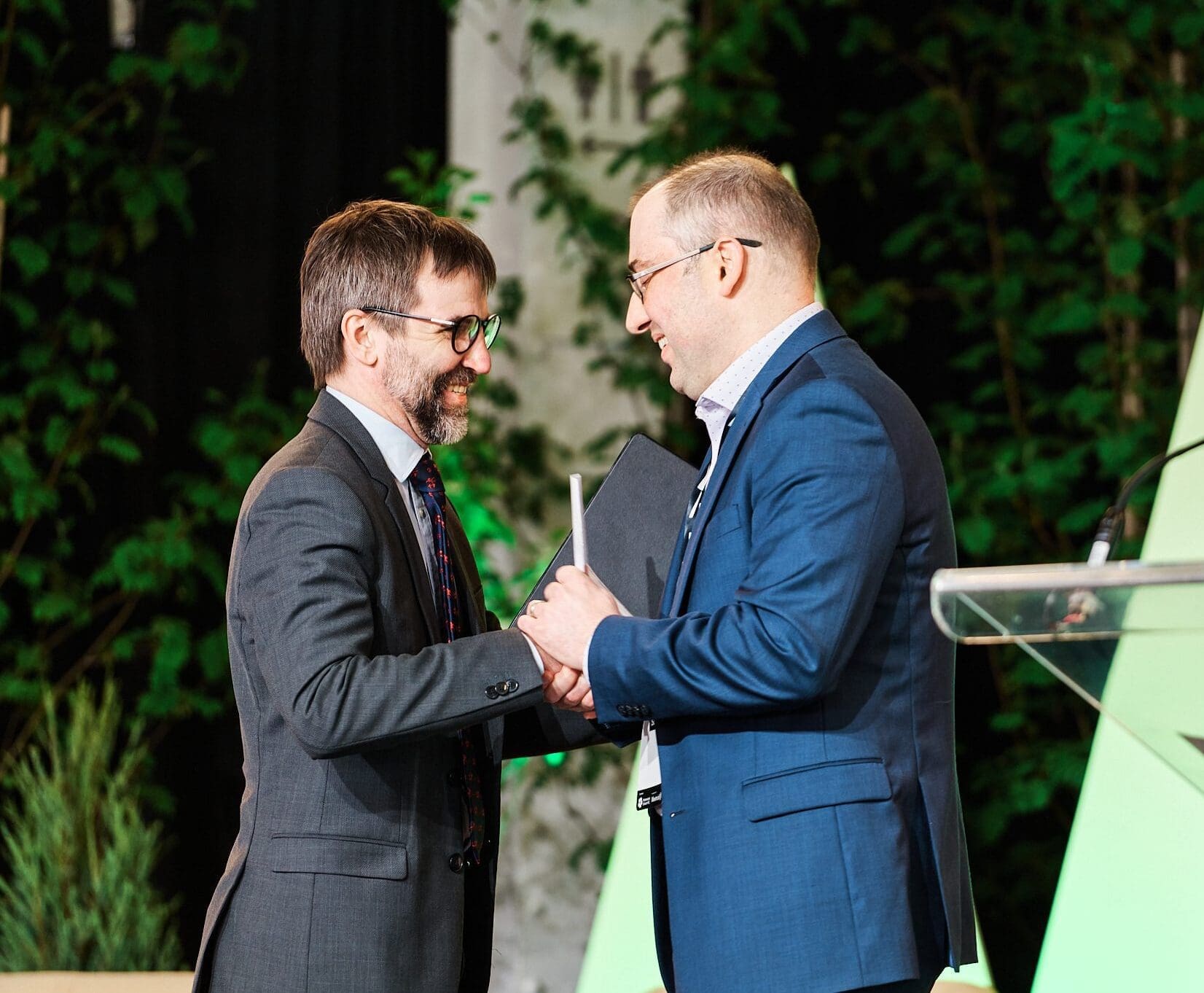 Le ministre canadien de l'environnement et du changement climatique, Steven Guilbeault, et le directeur du Fonds Climat du Grand Montréal, Vincent Moreau, sourient et se serrent la main sur scène. L'un d'eux tient un dossier alors qu'ils se tiennent au milieu de formes triangulaires et de feuillages éclairés en vert, à côté d'un podium transparent au Sommet Climat Montréal.