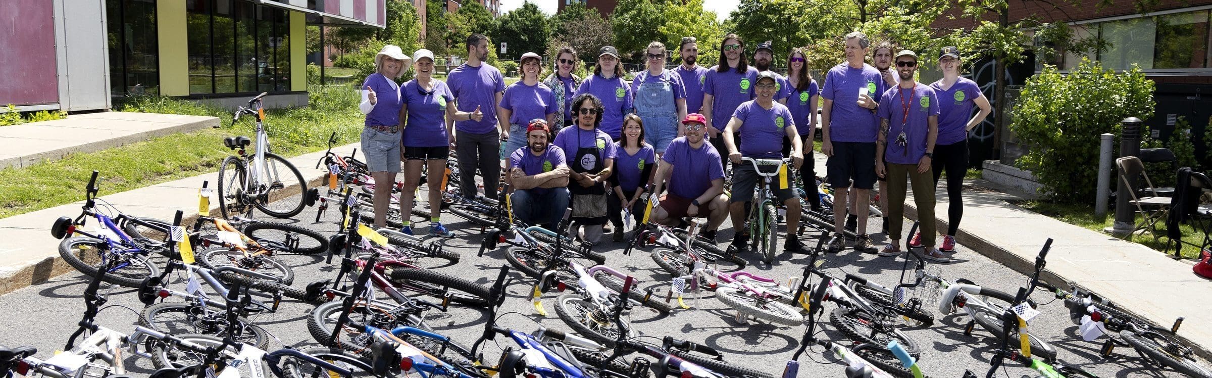 Un groupe de personnes portant des chemises violettes, faisant partie du projet Vélorution Couronne de Cyclo Nord-Sud, se rassemblent dans une rue avec des vélos d'enfants devant eux. Certains sont debout, d'autres accroupis, encadrés par des arbres et des bâtiments en arrière-plan.