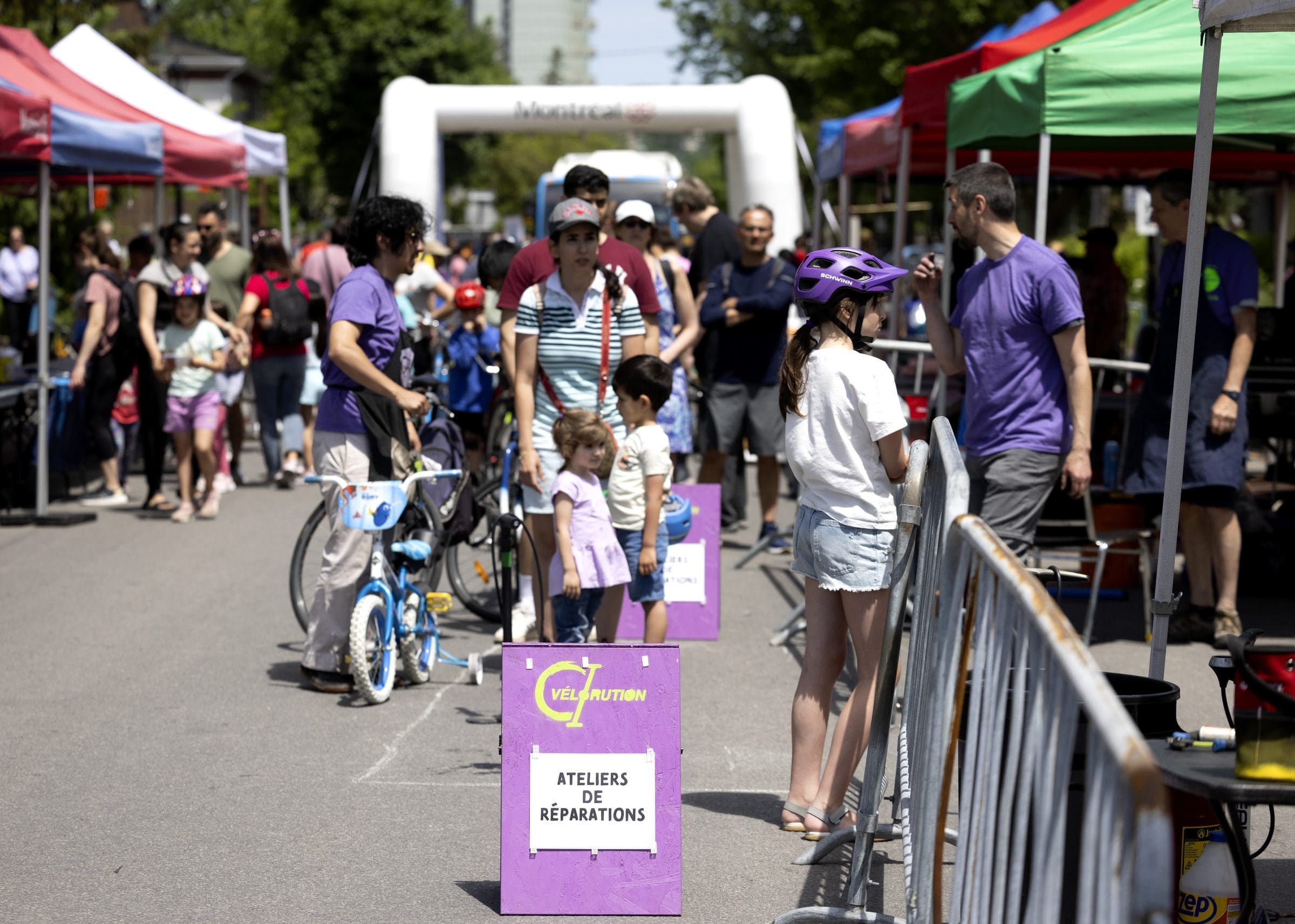 Lors d'un événement de réparation de vélos en plein air organisé par le projet Vélorution Couronne, des enfants et des adultes se rassemblent autour de vélos, entourés de tentes de réparation. Un panneau en français indique Ateliers de Réparations. La scène se déroule sur fond d'arbres et de ciel dégagé.