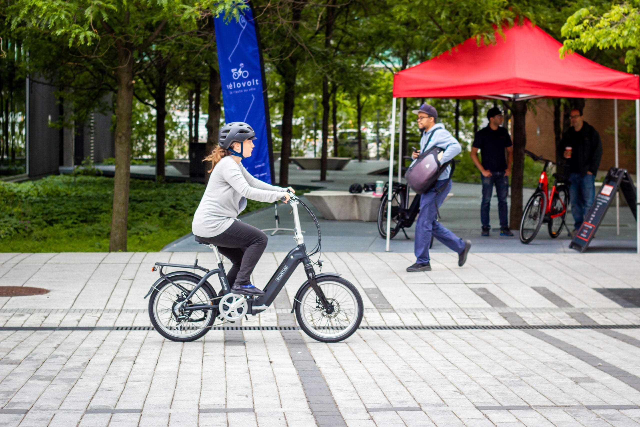 Une personne portant un casque passe à vélo devant une tente rouge, qui fait partie du projet Vélovolt d'Equiterre. À proximité, une autre personne marche en portant un grand sac, avec des arbres et une bannière bleue en arrière-plan, créant une scène énergique de place en plein air.