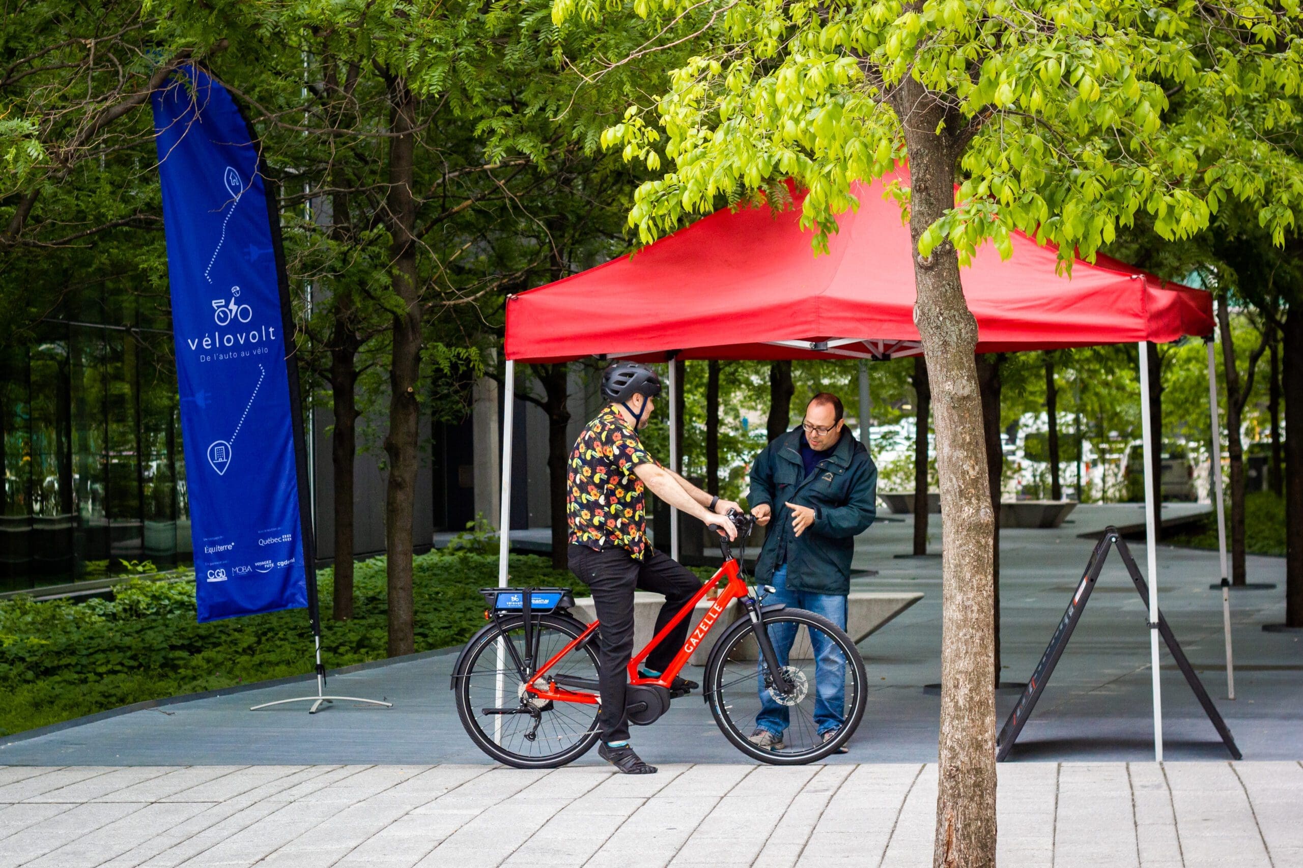 Une personne sur un vélo rouge discute sous une tente rouge, entourée d'arbres luxuriants. À proximité, un drapeau bleu avec vélovolt flotte, symbolisant le projet Vélovolt d'Équiterre. Le cycliste porte un casque et une chemise à motifs alors qu'ils discutent de transport durable.