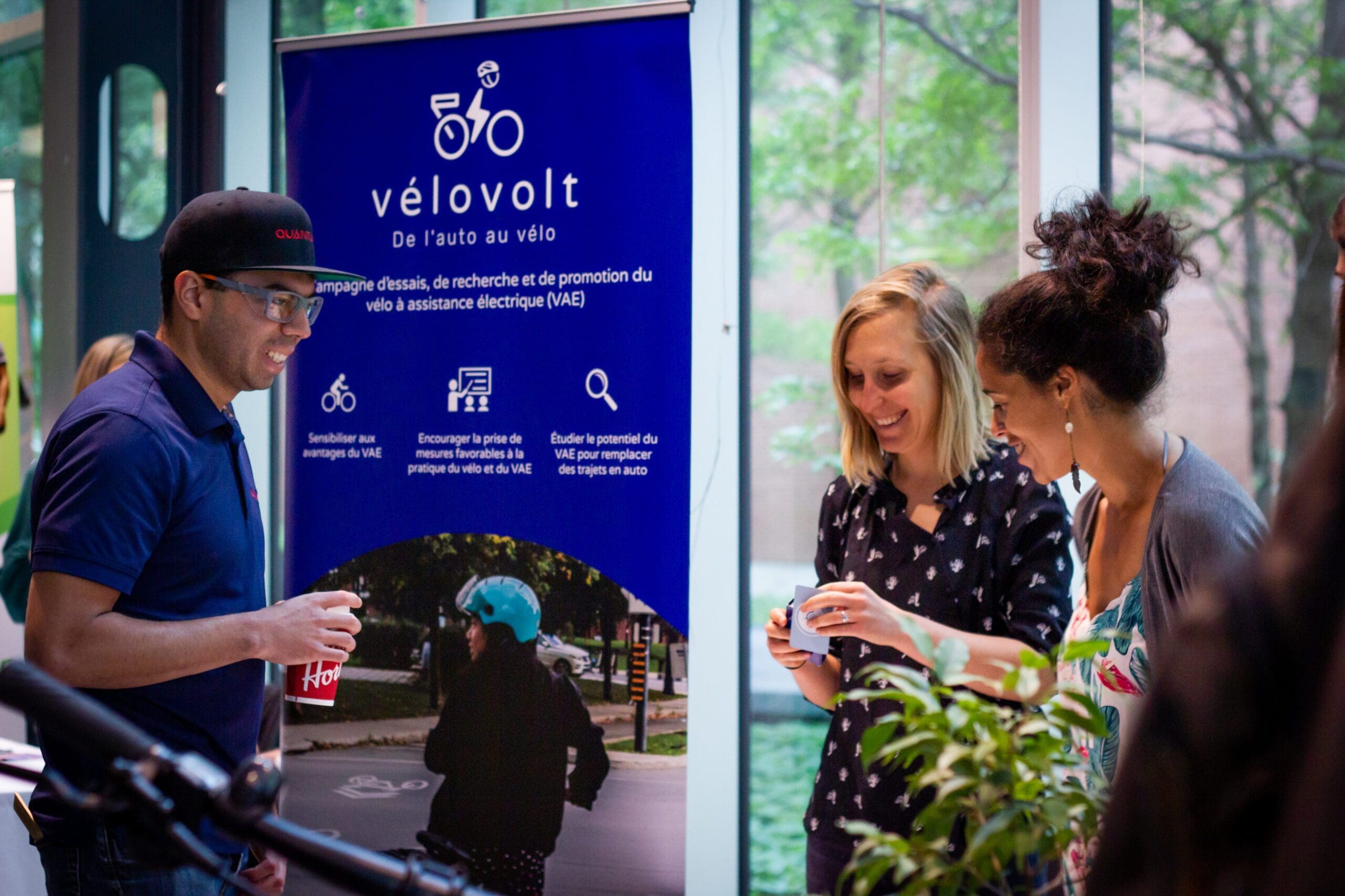 Trois personnes se tiennent devant une banderole de vélovolt en intérieur. Deux femmes regardent un téléphone en souriant, tandis qu'un homme portant une casquette tient un verre. La bannière présente des informations et des images relatives aux vélos électriques.