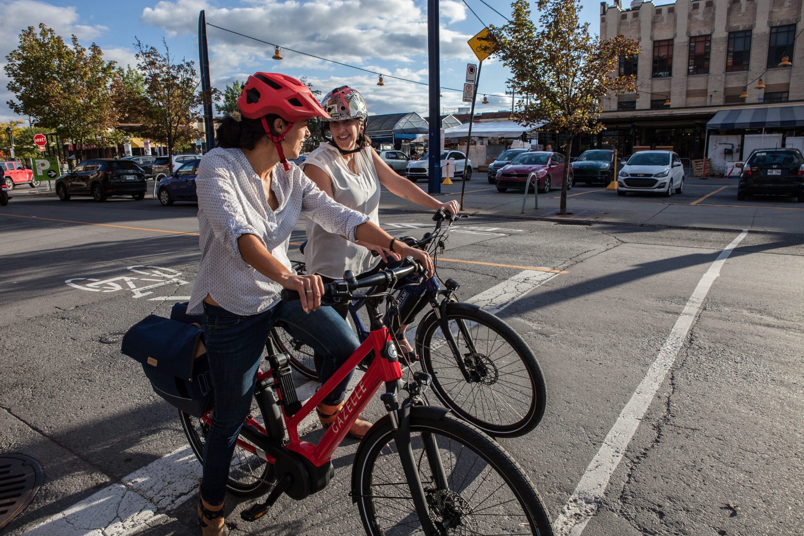 Deux personnes portant des casques sont assises sur des vélos à un passage piéton par une journée ensoleillée. Elles discutent et sourient. Des voitures sont garées le long de la rue et des arbres avec quelques feuilles d'automne bordent le trottoir. Un bâtiment avec des magasins se trouve à l'arrière-plan.