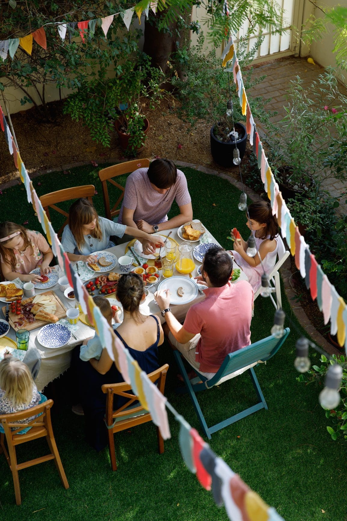 Un groupe de sept personnes est assis autour d'une table à manger en plein air, décorée de guirlandes colorées. La table est chargée de divers plats. Elles sont situées sur une pelouse entourée d'arbres et de plantes. La scène évoque une réunion festive dans l'arrière-cour.