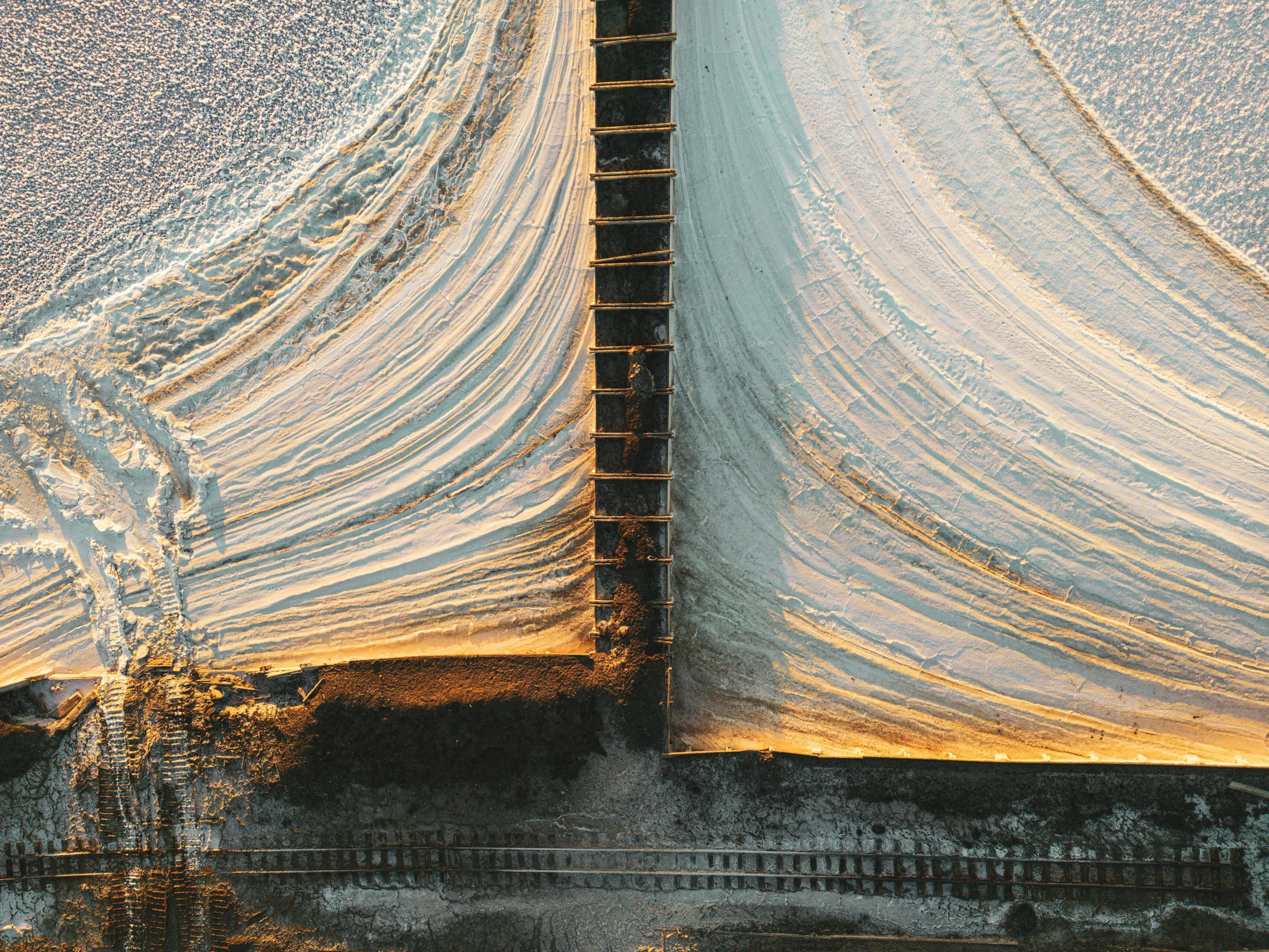 Vue aérienne de deux grands bassins d'évaporation du sel. Les bassins sont divisés par une clôture ou une passerelle en bois, créant des motifs géométriques dans les tons blanc et beige clair. Des traces de pneus et des empreintes de pas sont visibles en bas à gauche.