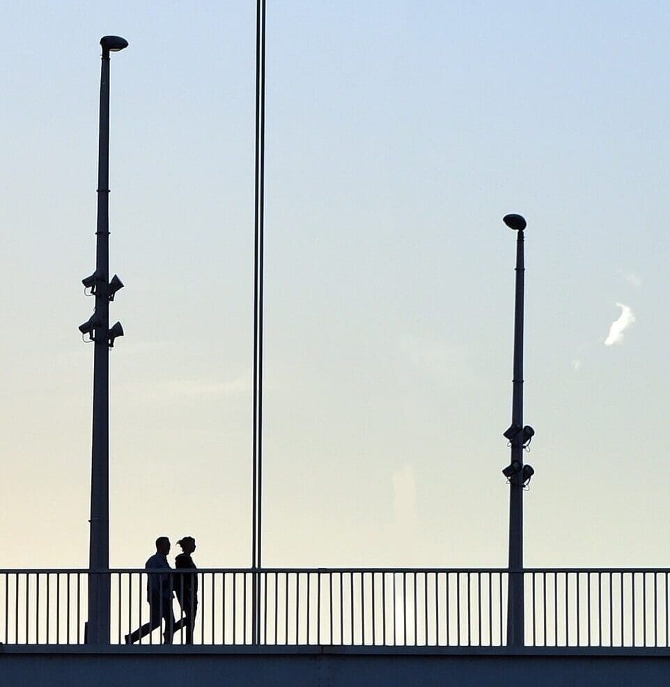 Silhouettes de deux personnes traversant un pont au coucher du soleil. Les câbles du pont et les lampadaires sont visibles dans un ciel clair avec une lueur chaude près de l'horizon.