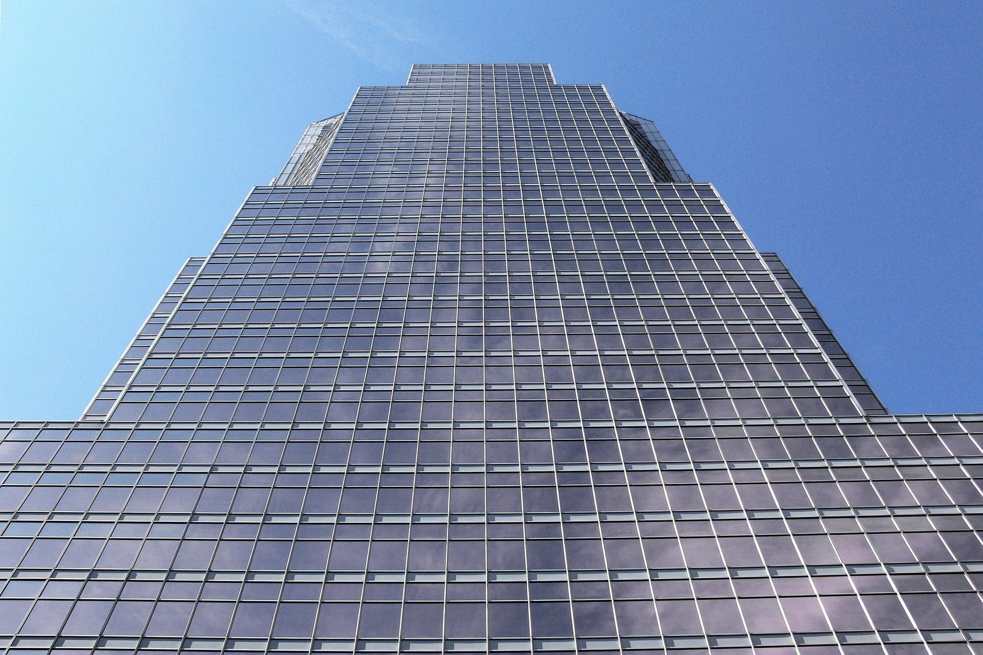 Vue plongeante sur un gratte-ciel de grande hauteur avec une grille de fenêtres en verre réfléchissant sur un ciel bleu clair. Le bâtiment s'amincit légèrement à mesure qu'il s'élève, mettant en valeur une conception architecturale moderne.