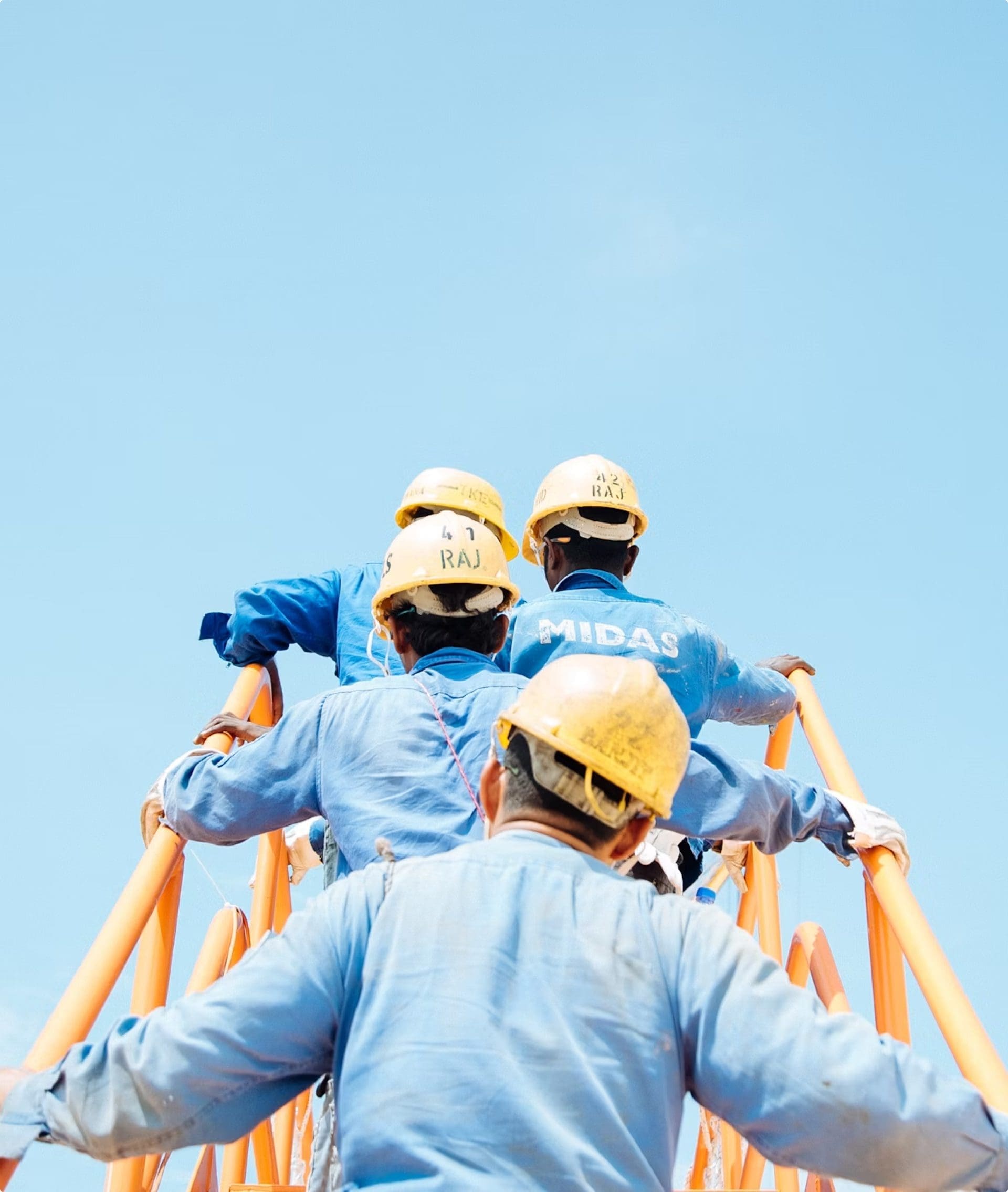 Des ouvriers en uniformes bleus et casques de chantier jaunes escaladent une structure métallique orange sur fond de ciel bleu clair. Le mot « Midas » est visible au dos de la chemise d'un ouvrier.