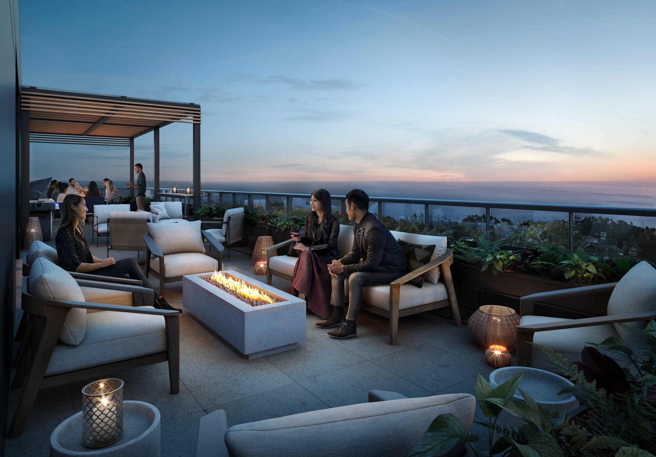 Des personnes sont assises sur un patio sur le toit au coucher du soleil, rassemblées autour d'un foyer rectangulaire. Les condos de Courcelette offrent un mobilier d'extérieur confortable, avec une vue sur le paysage urbain et le ciel en arrière-plan. Des lanternes et des plantes ajoutent une touche décorative.