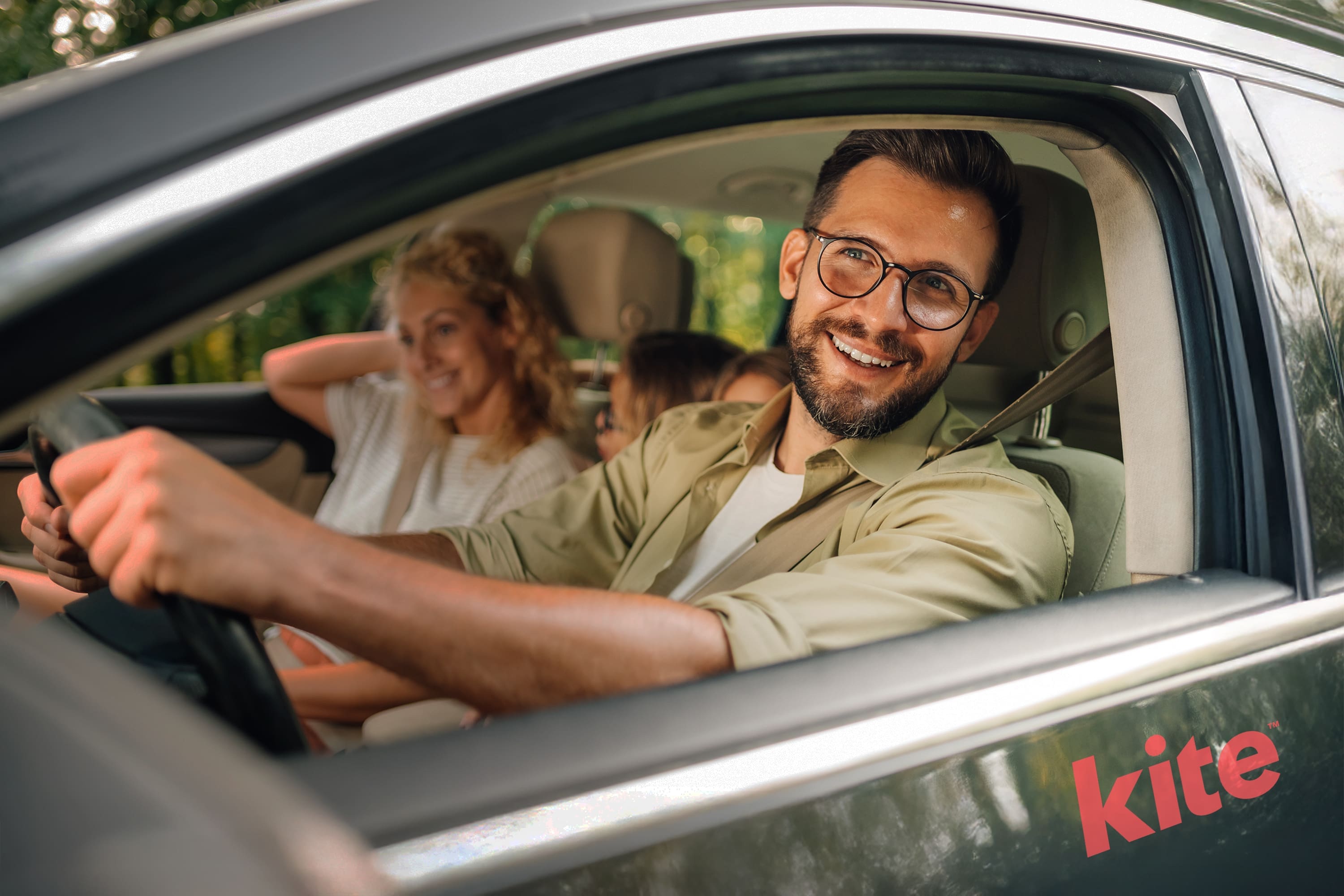 Un homme portant des lunettes sourit chaleureusement depuis le siège du conducteur d'une voiture portant l'étiquette cerf-volant. Derrière lui, une femme et un enfant partagent la joie, leurs sourires éclatants se détachant sur un arrière-plan de feuillage vert et luxuriant légèrement flou.