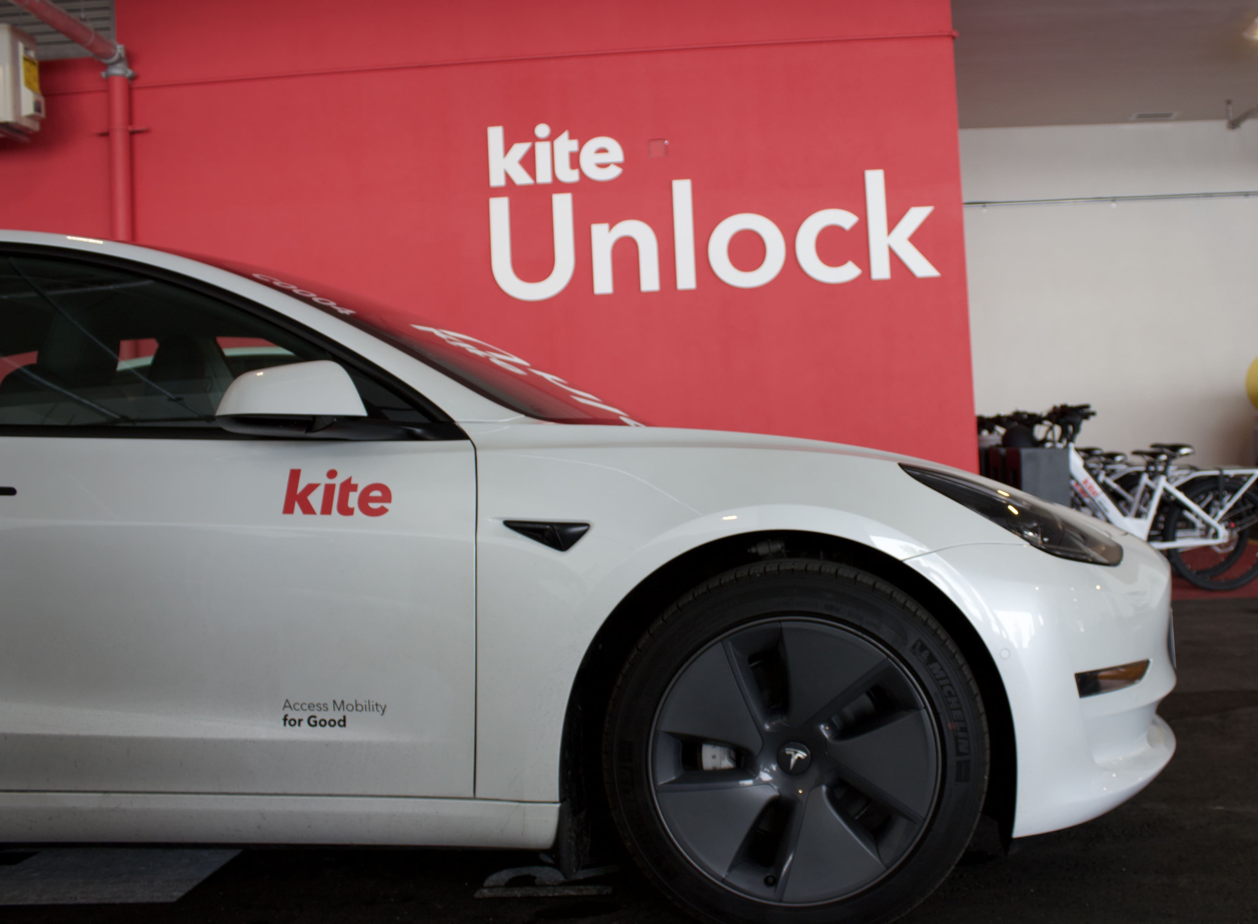 Une voiture blanche portant l'inscription Kite sur son flanc est garée à l'intérieur, contre un mur rouge frappant portant l'inscription Kite Unlock en lettres blanches en gras. Des bicyclettes se tiennent tranquillement à l'arrière-plan, complétant la scène.