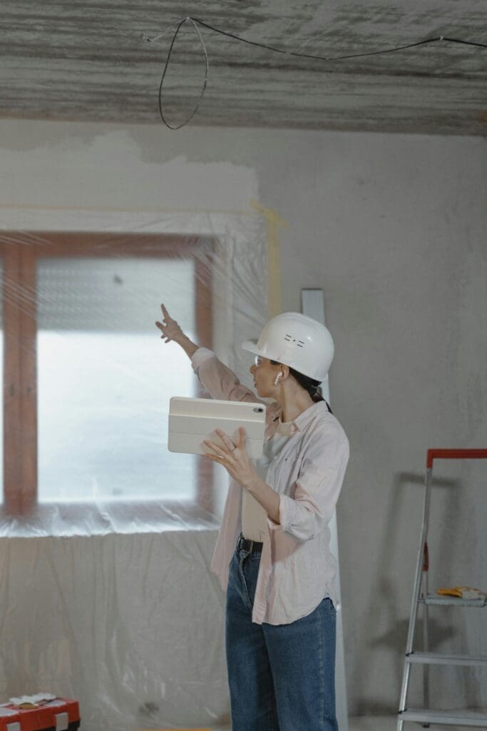 Une femme porte un casque de chantier et des documents de travaux en main dans une pièce en travaux